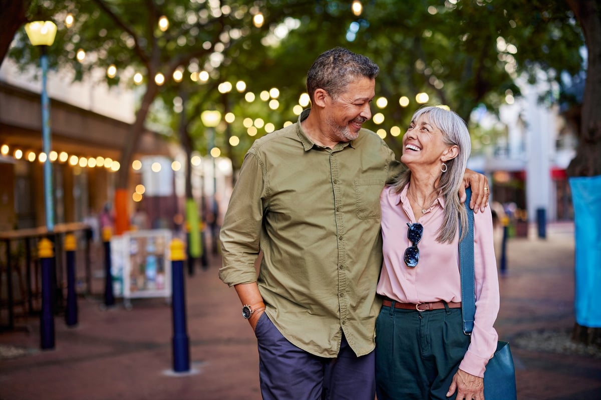 Happy multiethnic man and smiling woman enjoy a romantic evening outdoors. Mature couple shares a loving moment while walking in a european city. Smiling senior couple walks closely together under string lights with copy space.