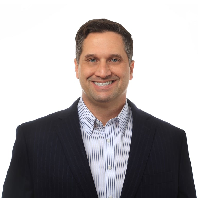 Professional headshot of a smiling man wearing a black blazer and light blue striped shirt against a white background