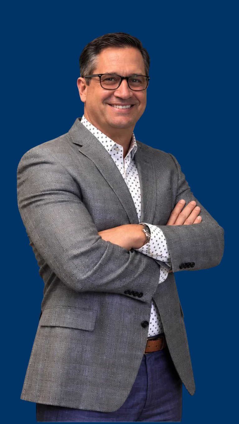 Smiling man in gray blazer and glasses with arms crossed, standing against a blue background